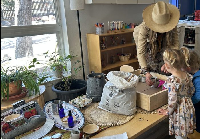 A teacher in a wide-brimmed hat shows a young child a project at a sun-lit table with plants and natural materials