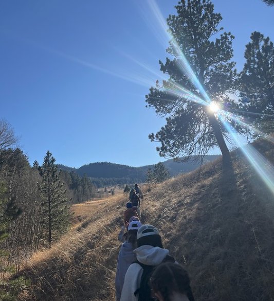A line of children hiking on a sunny mountain trail with foothills behind them