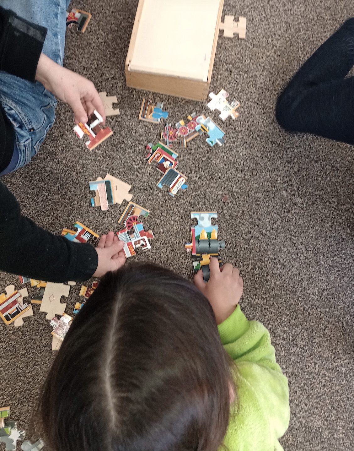 A child working a colorful train puzzle on the carpet alongside an adult helper